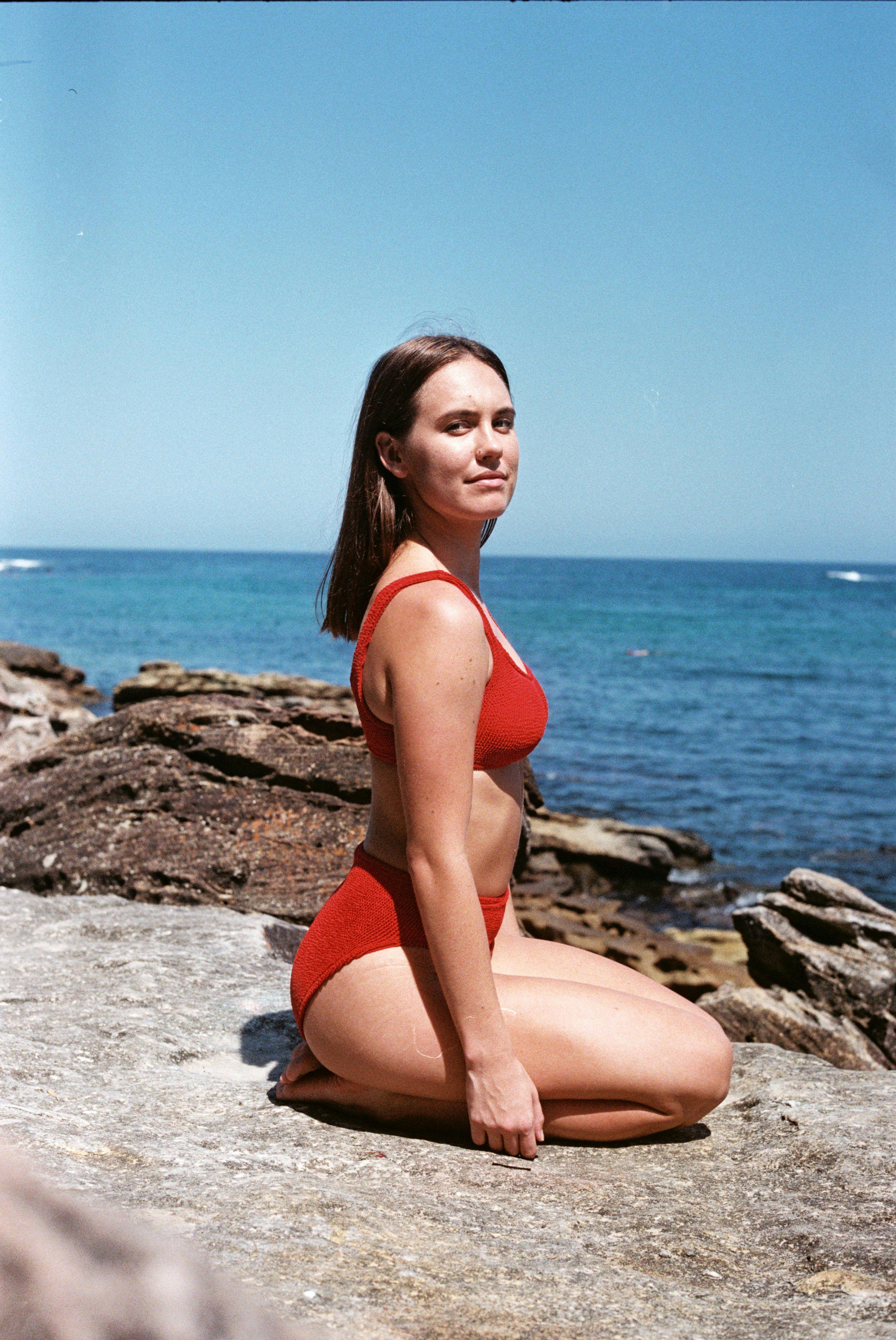 Marlee kneeling on rocks by the ocean in red bikini